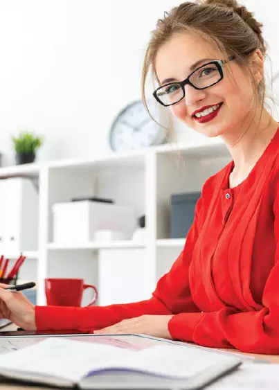A woman in glasses and a red blouse smiles at her desk with an open book, red mug, and pens — representing Leading Digital success.
